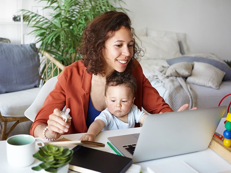 A mother holding her baby while staying involved in community matters on her computer. Positive results of digital government engagement and communications.