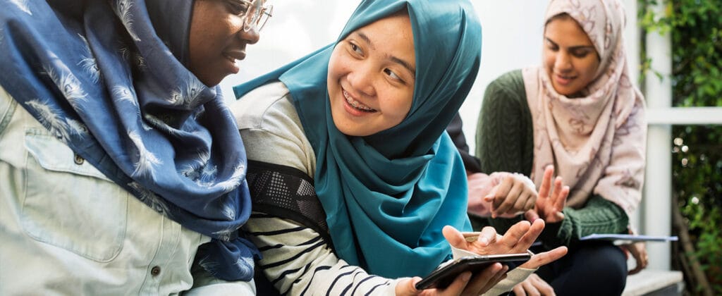 Three young women sitting while two use a digital device