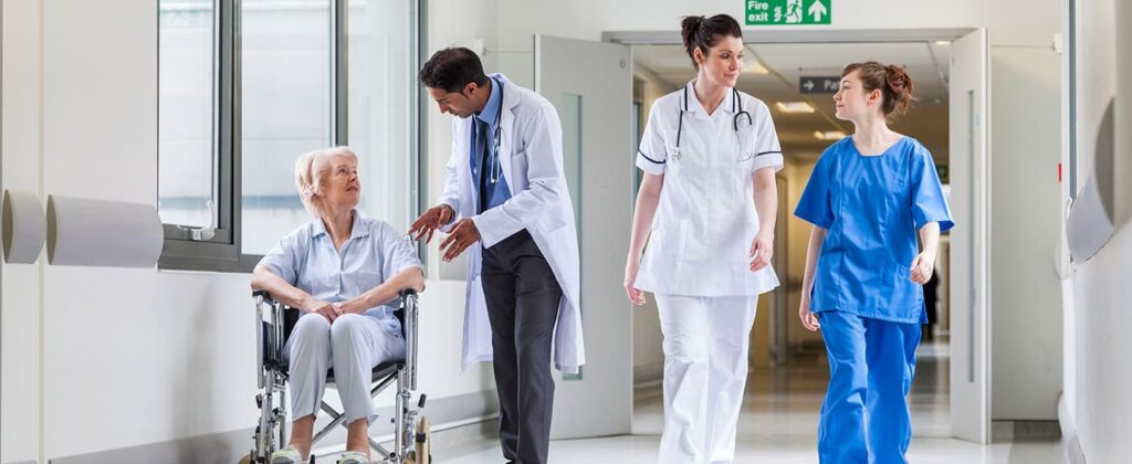 Various hospital staff walking down corridor while a doctor speaks to a senior woman in a wheelchair